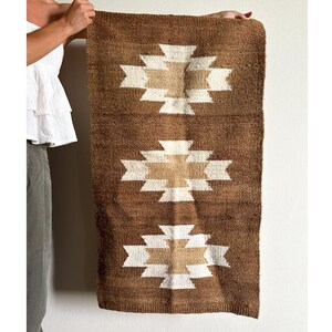May include: Brown and beige woven wall hanging with a geometric pattern. The rectangular rug features three repeating white and beige geometric designs against a brown background. The rug is held up by a person.