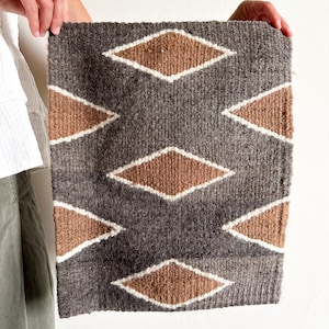Puede incluir: Textil rectangular tejido con un patrón de diamantes repetido. Los diamantes son marrones con bordes blancos, sobre un fondo gris. El textil es sostenido por una persona.