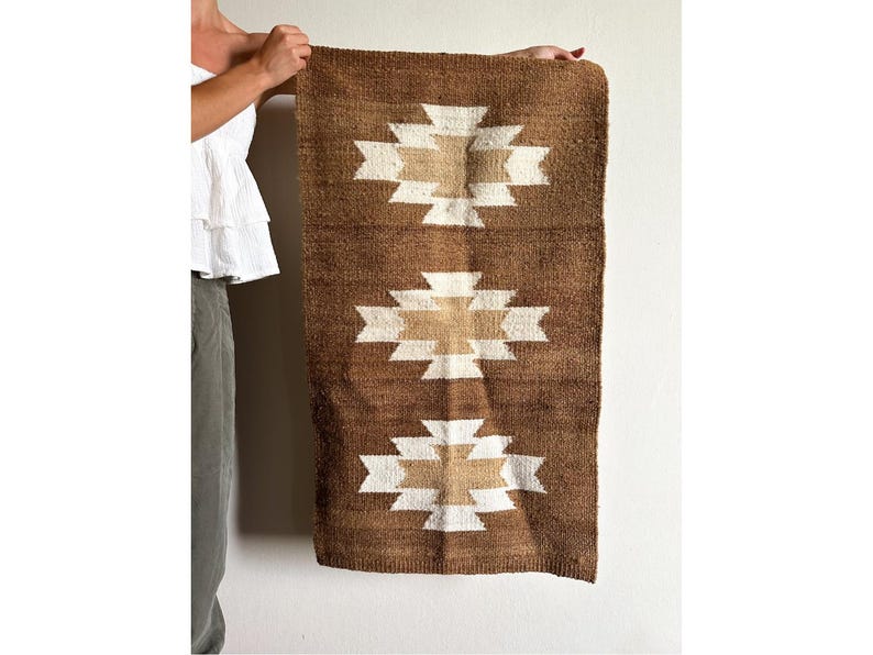 May include: Brown and beige woven wall hanging with a geometric pattern. The rectangular rug features three repeating white and beige geometric designs against a brown background. The rug is held up by a person.