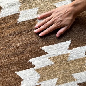 May include: Close-up of a brown and white patterned rug. The rug features a geometric design with white and tan shapes against a brown background. The texture of the rug is visible, and a hand is resting on the surface.