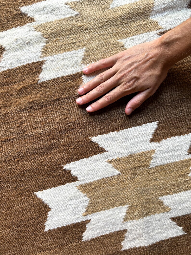 May include: Close-up of a brown and white patterned rug. The rug features a geometric design with white and tan shapes against a brown background. The texture of the rug is visible, and a hand is resting on the surface.