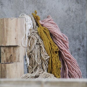 May include: A collection of colorful yarn skeins in shades of white, tan, mustard yellow, and dusty rose. The yarn is thick and textured, resting against a weathered wooden surface and a gray concrete wall.