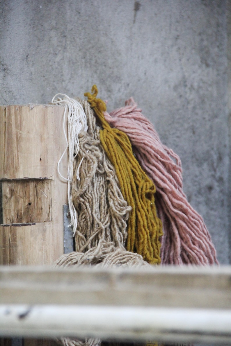 May include: A collection of colorful yarn skeins in shades of white, tan, mustard yellow, and dusty rose. The yarn is thick and textured, resting against a weathered wooden surface and a gray concrete wall.