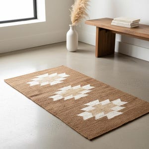 May include: A rectangular brown rug with three repeating geometric patterns in white and tan. The rug is made of a woven material and is placed on a concrete floor. The rug is a decorative element for a living space.