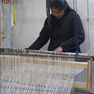 May include: A woman weaving on a wooden loom. The loom is filled with vertical white threads, and the woman is wearing a black jacket. Various tools and materials hang nearby, suggesting a craft workshop.