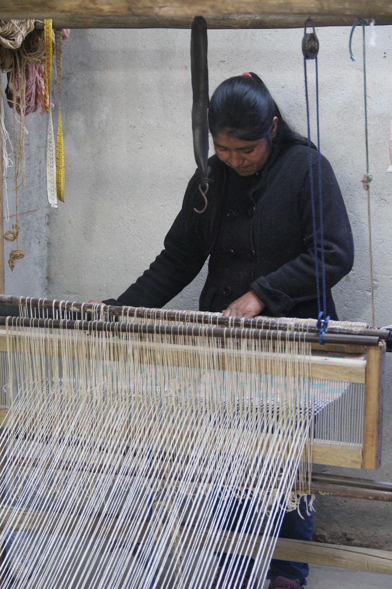 May include: A woman weaving on a wooden loom. The loom is filled with vertical white threads, and the woman is wearing a black jacket. Various tools and materials hang nearby, suggesting a craft workshop.
