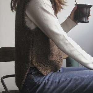 Puede incluir: Un chaleco de punto marrón se usa sobre una camisa blanca de manga larga. La persona sostiene una taza de cuero marrón oscuro con una pajita de metal. La persona lleva vaqueros azules y está sentada en una silla de madera.
