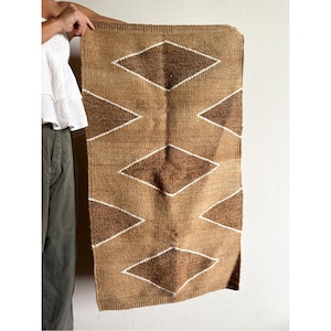 Puede incluir: Una alfombra rectangular tejida con un patrón geométrico de diamantes. La alfombra presenta diamantes marrones y beige alternados, delineados en blanco. La alfombra se sostiene contra una pared blanca, mostrando su textura y diseño.