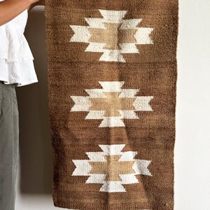 May include: Brown and beige woven wall hanging with a geometric pattern. The rectangular textile features three repeating designs of a white and beige cross-like shape on a brown background. The wall hanging is made of a textured material.