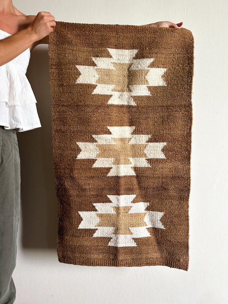 May include: Brown and beige woven wall hanging with a geometric pattern. The rectangular textile features three repeating designs of a white and beige cross-like shape on a brown background. The wall hanging is made of a textured material.