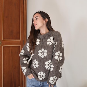 Puede incluir: Un suéter marrón con patrones florales blancos. El suéter tiene cuello redondo y mangas largas. La persona lleva vaqueros azules. El suéter está hecho de un material grueso y tejido.