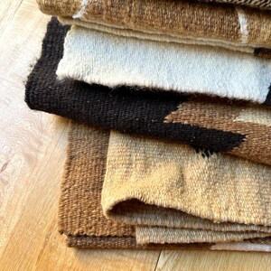 May include: A stack of woven textiles in various earth tones. The textiles include shades of brown, beige, and black, with some featuring geometric patterns and white accents. The fabrics appear to be thick and textured, suggesting a rustic or bohemian style.