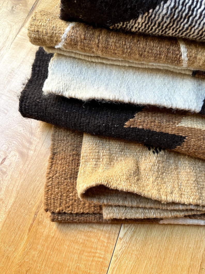 May include: A stack of woven textiles in various earth tones. The textiles include shades of brown, beige, and black, with some featuring geometric patterns and white accents. The fabrics appear to be thick and textured, suggesting a rustic or bohemian style.