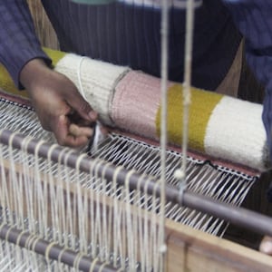 May include: A person weaving a colorful textile on a loom. The textile features sections of white, pink, and yellow. The loom is made of wood and metal, with numerous vertical threads. The person's hands are visible as they work on the weaving process.
