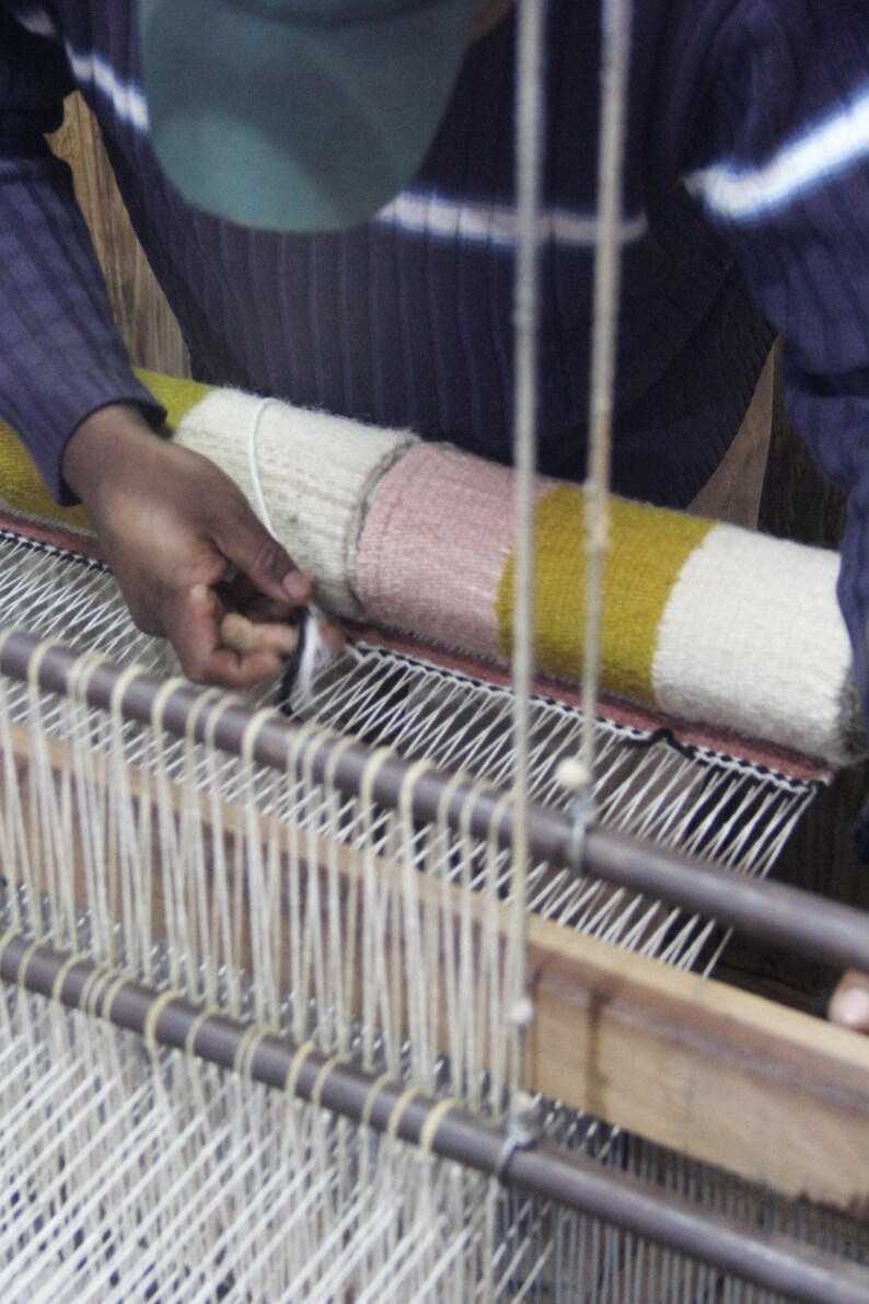 May include: A person weaving a colorful textile on a loom. The textile features sections of white, pink, and yellow. The loom is made of wood and metal, with numerous vertical threads. The person's hands are visible as they work on the weaving process.