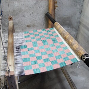 May include: A wooden weaving loom with a partially completed textile. The textile features a checkered pattern in shades of pink and teal. The loom is set against a gray concrete wall, with measuring tapes hanging nearby.