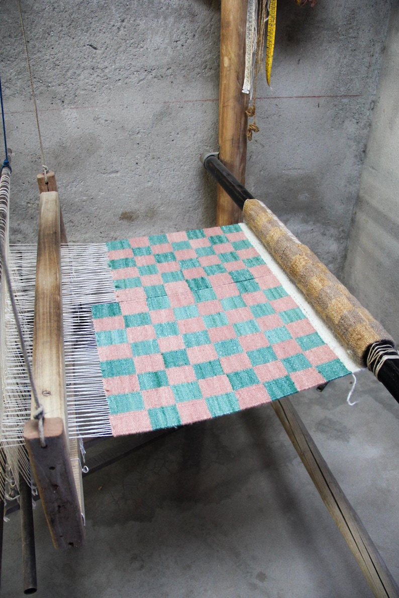 May include: A wooden weaving loom with a partially completed textile. The textile features a checkered pattern in shades of pink and teal. The loom is set against a gray concrete wall, with measuring tapes hanging nearby.