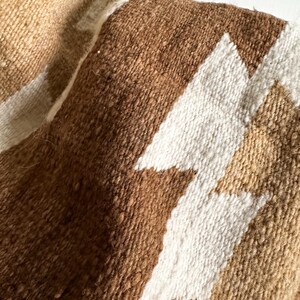 May include: Close-up of a woven textile with a geometric pattern. The fabric features a textured surface with shades of brown, tan, and white. The design includes angular shapes, creating a visual contrast.
