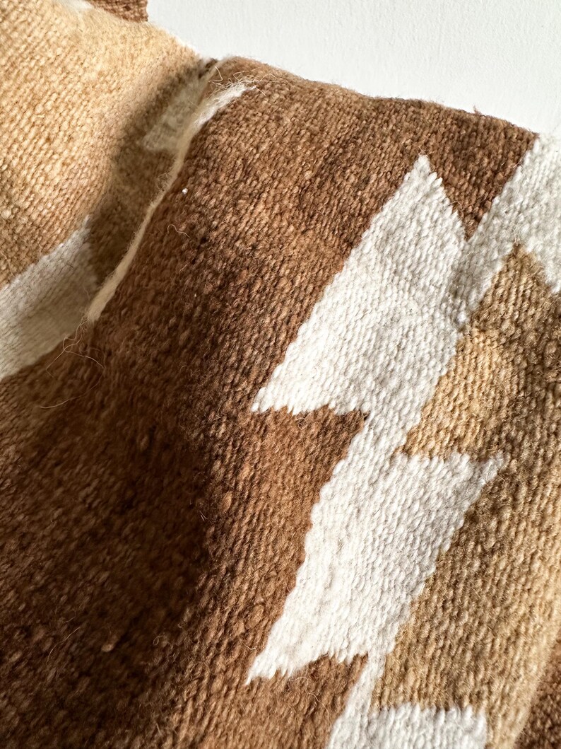 May include: Close-up of a woven textile with a geometric pattern. The fabric features a textured surface with shades of brown, tan, and white. The design includes angular shapes, creating a visual contrast.