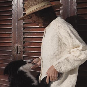 Puede incluir: Una mujer con una rebeca de punto color crema y un sombrero de paja con una cinta beige. Interactúa con un perro blanco y negro. El fondo es una puerta de madera marrón oscuro.
