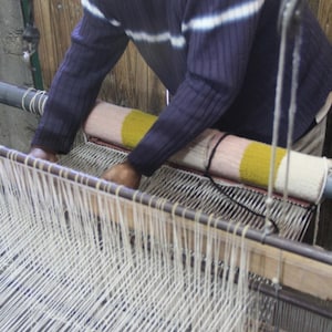 May include: A person weaving on a loom. The person is wearing a blue sweater with white stripes. The loom has white threads and a colorful woven piece. The background is a wooden wall.