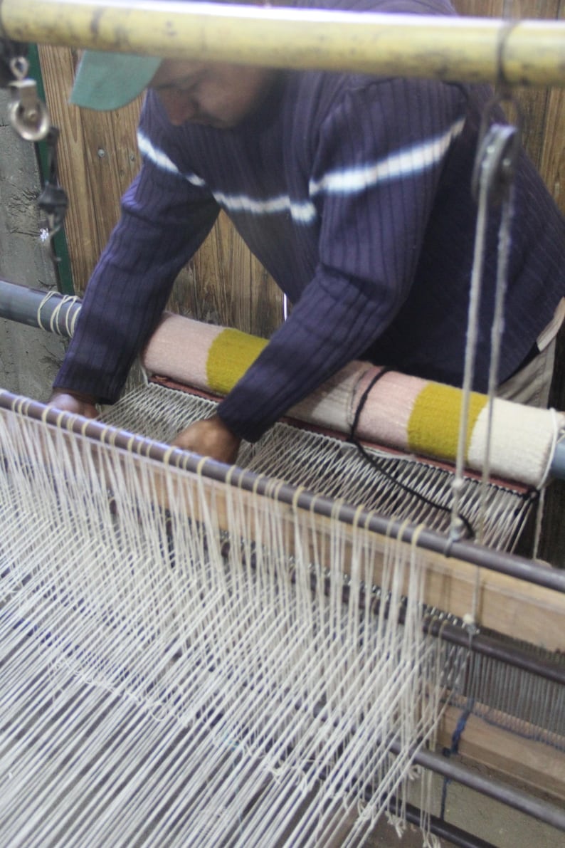 May include: A person weaving on a loom. The person is wearing a blue sweater with white stripes. The loom has white threads and a colorful woven piece. The background is a wooden wall.