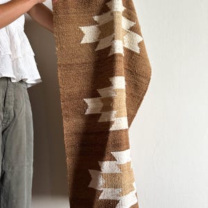 May include: A brown woven textile with a geometric pattern in white and tan. The textile is rectangular and appears to be a rug or wall hanging. The design features repeating shapes. The textile is being held up against a white wall.
