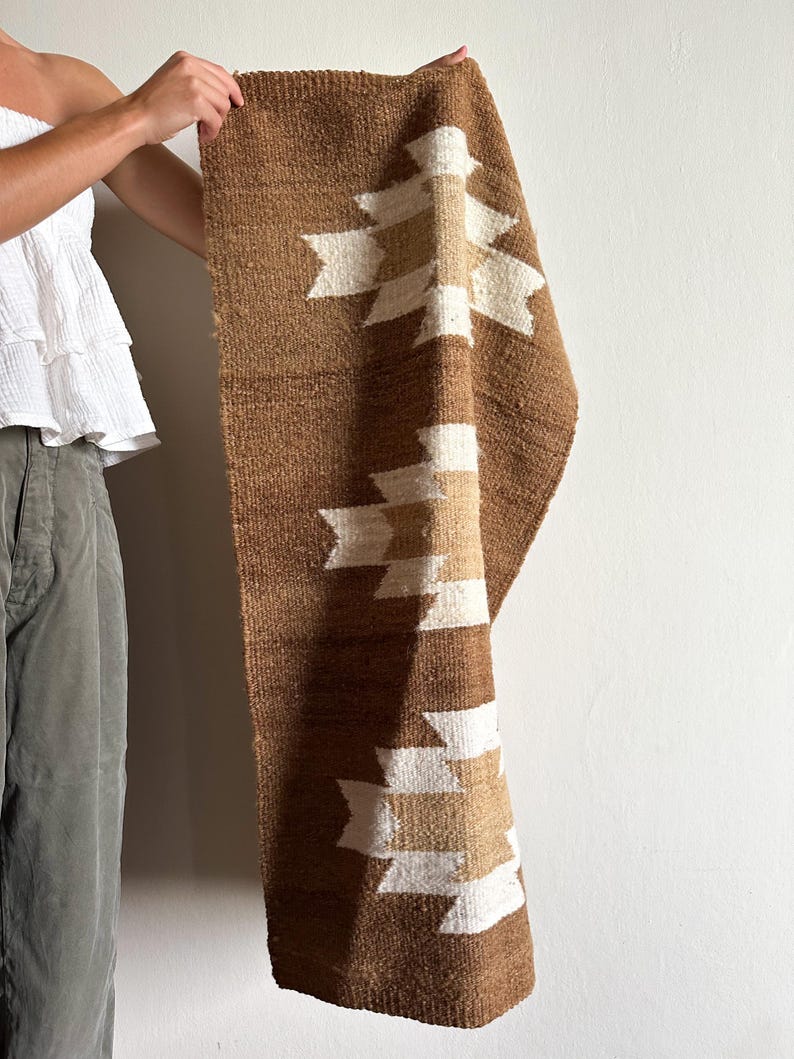 May include: A brown woven textile with a geometric pattern in white and tan. The textile is rectangular and appears to be a rug or wall hanging. The design features repeating shapes. The textile is being held up against a white wall.
