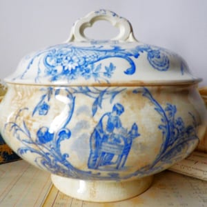 May include: A cream-colored ceramic tureen with a matching lid and two side handles. The exterior features blue transferware patterns depicting floral motifs and a figure working at a table, with visible brown crazing across the glazed surface.