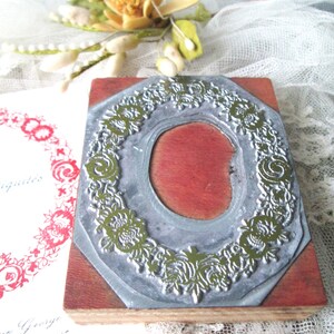 May include: A vintage wooden stamp with a floral wreath design. The stamp has a silver metal frame and a red interior. The stamp is on a wooden base. A printed card with red floral design is visible.