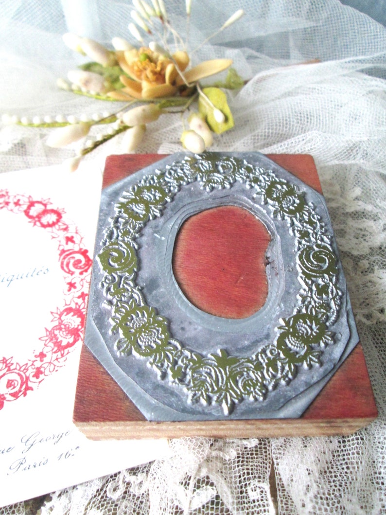May include: A vintage wooden stamp with a floral wreath design. The stamp has a silver metal frame and a red interior. The stamp is on a wooden base. A printed card with red floral design is visible.