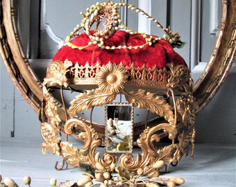 May include: A gold and red crown with a velvet cushion and a small mirror. The crown is decorated with ornate details and floral motifs.