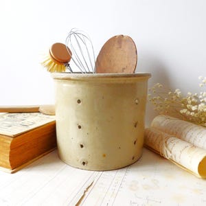 Op de afbeelding: Een cilindrische beige keramische pot met een reeks kleine, gelijkmatig verdeelde ronde gaten aan de zijkant. De pot bevat een houten afwasborstel, een metalen garde en een ronde houten schijf, met een dik boek en papier in de buurt.