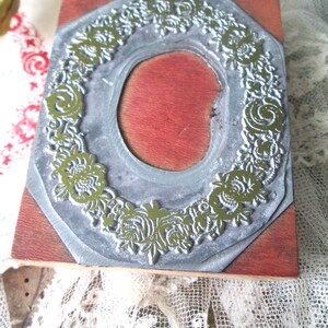May include: A vintage printing block with a floral design. The block features a silver-colored metal frame with a detailed green floral pattern surrounding a red oval center. The block is mounted on a wooden base.