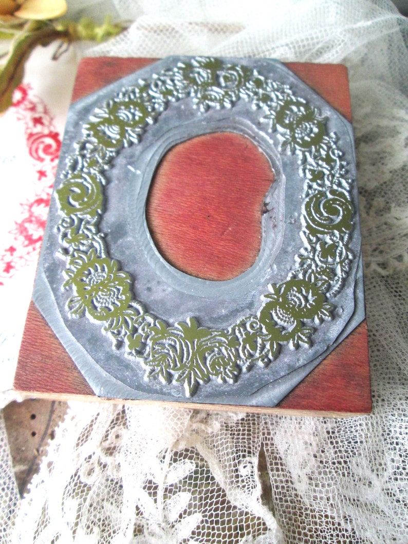 May include: A vintage printing block with a floral design. The block features a silver-colored metal frame with a detailed green floral pattern surrounding a red oval center. The block is mounted on a wooden base.