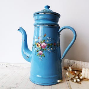 Op de afbeelding: Een lichtblauwe emaille koffiepot met een bloemmotief van roze rozen, gele viooltjes en groene bladeren. De cilindrische pot heeft een bijpassend deksel, een gebogen handvat en een tuit, met zichtbare slijtage langs de randen van het metaal.