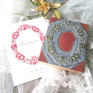 May include: A wooden stamp with a floral design and a red ink pad, next to a card with the text "Romantiquit&eacute;s" and a red floral wreath. The items are on a white lace surface.