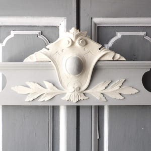 May include: A decorative architectural element in shades of gray and white. The design features a central oval shape, flanked by stylized leaves and scrollwork, mounted on a gray panel. The background is a gray wooden door.