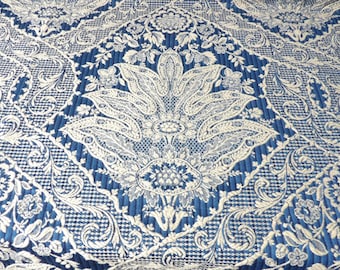 May include: Close-up of a blue and white patterned fabric. The design features a large, central floral motif surrounded by intricate scrollwork and geometric patterns. The fabric has a textured appearance.