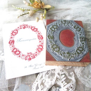 May include: A vintage-style stamp with a floral design and a red wooden base. A card with the word "Romantiquit&eacute;s" in script and a floral wreath is next to the stamp. The background includes lace and a small floral arrangement.