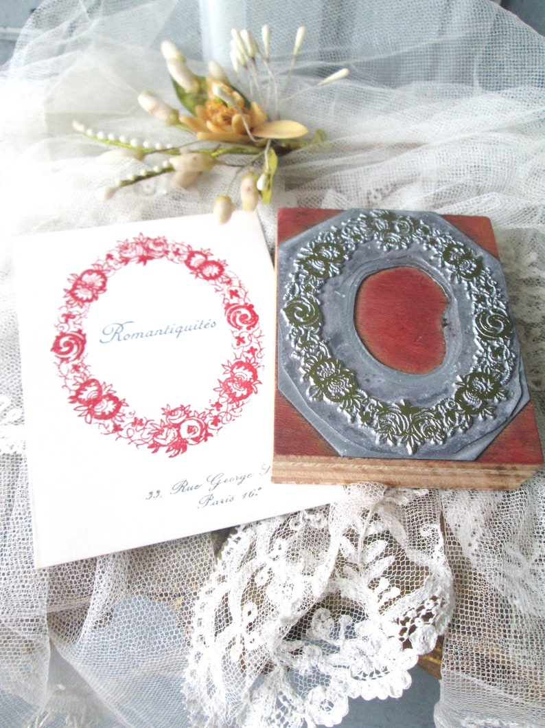 May include: A vintage-style stamp with a floral design and a red wooden base. A card with the word "Romantiquit&eacute;s" in script and a floral wreath is next to the stamp. The background includes lace and a small floral arrangement.