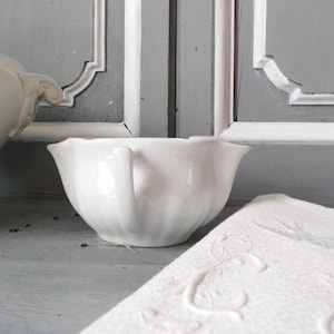 Peut inclure: Un bol en porcelaine blanc avec une anse et un bord festonné. Un tissu blanc à motifs est partiellement visible à droite. Le bol est posé devant un fond d'armoire grise.