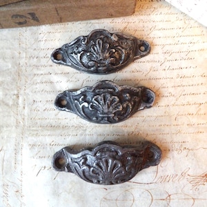 Op de afbeelding: Drie antieke zilverkleurige ladegrepen met sierlijke bloemmotieven. De grepen zijn halfrond en hebben een verhoogd centraal motief.