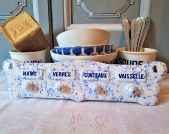 May include: A white ceramic dish rack with blue and white speckled glaze. The rack has four compartments labeled "Mains", "Verres", "Couteaux", and "Vaisselle".