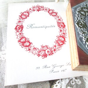 May include: A white card with a red floral wreath design and the word "Romantiquit&eacute;s" in a script font. The card also includes the address "33. Rue George Sand Paris 16.". A wooden stamp is visible on the right.