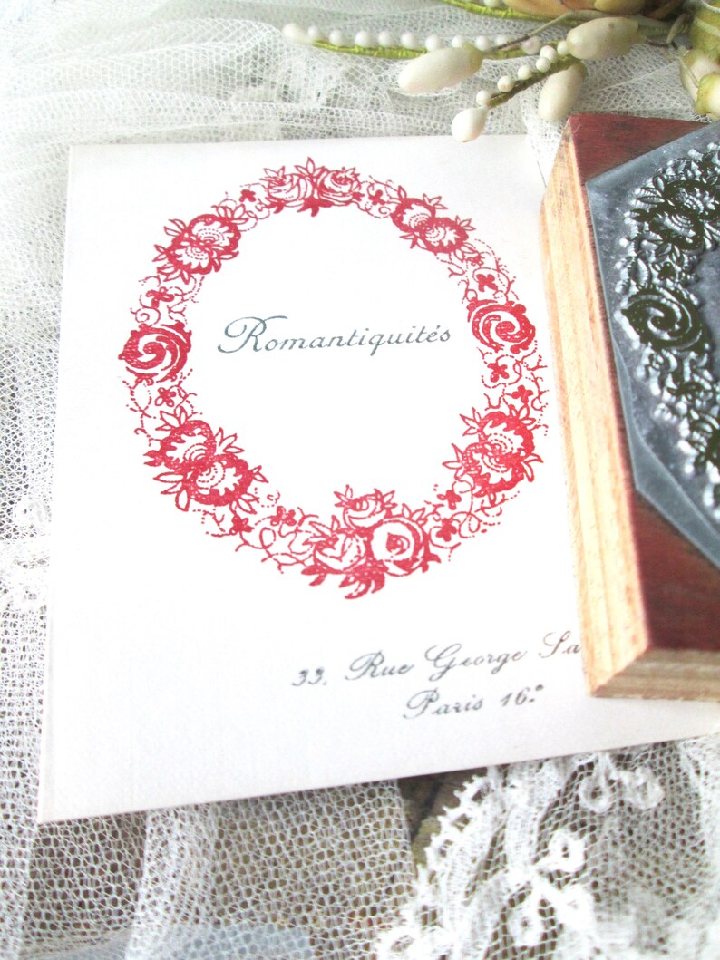 May include: A white card with a red floral wreath design and the word "Romantiquit&eacute;s" in a script font. The card also includes the address "33. Rue George Sand Paris 16.". A wooden stamp is visible on the right.