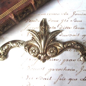 Peut inclure: Une ornement métallique décoratif avec un motif floral. L'ornement est en laiton et a une finition dorée. Il présente un motif central de fleur de lys avec deux motifs floraux plus petits de chaque côté. L'ornement est conçu pour être fixé à une surface, comme un meuble ou un mur.