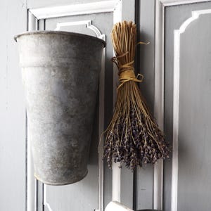 May include: A grey metal bucket hangs on a white and grey panelled wall. A bundle of dried lavender is hanging next to the bucket.