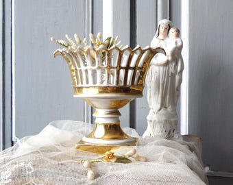 May include: A white and gold porcelain centerpiece with a decorative openwork design, next to a white statue of Mary holding baby Jesus. The centerpiece sits on a lace cloth, with a light gray background.
