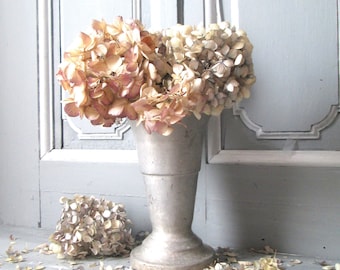 May include: A silver vase with a bouquet of dried hydrangeas in shades of pink and white. The flowers are arranged in a loose, natural style. The vase is sitting on a light gray surface.